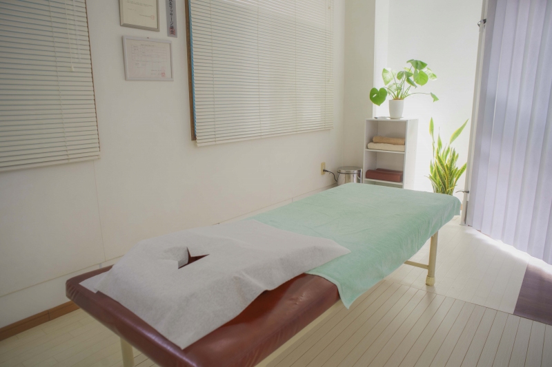 リラクゼーションと痛みの治療が共存する快晴鍼灸院の施術室 快晴鍼灸院の院内の様子。白い床と壁に自然光が差し込む施術室に、施術ベッドと観葉植物、タオル棚を配置したリラックスできる空間。リラクゼーションから首こり・肩こり・緊張型頭痛などの鍼灸治療まで対応可能な、開放感と落ち着きを兼ね備えた施術スペース。