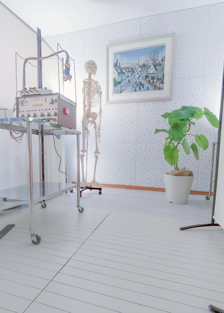 快晴鍼灸院のメディカルギャラリーゾーン(多目的ユーティリティスペース) 足柄上郡開成町・快晴鍼灸院の白を基調とした多目的ゾーン。白を基調としたメディカルギャラリーゾーンに、骨格模型と医療機器、観葉植物、壁のアートを配置し、首こり・緊張型頭痛など筋・筋膜性疼痛に取り組む専門性と、やわらかな院内の雰囲気を同時に表現している。問診スペース・応接室・院内ギャラリーを兼ねるユーティリティスペース。